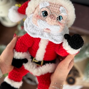 Pode incluir: Boneca de Papai Noel de croch&ecirc;, vestida de vermelho e branco com cinto e luvas pretas. A boneca tem uma barba branca fofa, um chap&eacute;u vermelho e &oacute;culos com arma&ccedil;&atilde;o de arame. Uma decora&ccedil;&atilde;o de Natal encantadora.