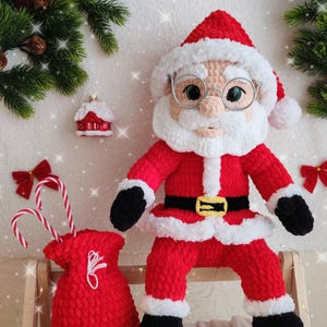 Pode incluir: Boneca de croch&ecirc; feita &agrave; m&atilde;o do Papai Noel, vestindo um terno vermelho com acabamento branco, um cinto preto com fivela dourada e botas pretas. A boneca tem barba branca, &oacute;culos e um chap&eacute;u vermelho. Uma sacola vermelha com bengalas doces est&aacute; por perto.