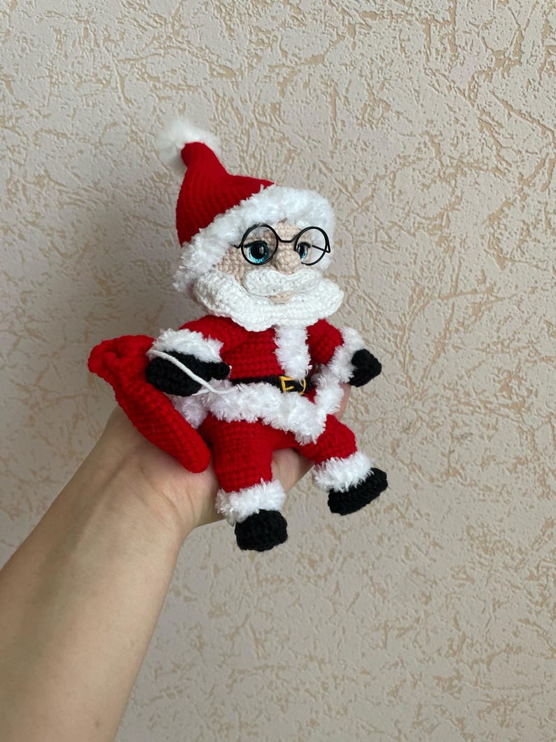 Pode incluir: Boneca de croch&ecirc; do Papai Noel, com roupa e chap&eacute;u vermelhos, barba e acabamento brancos. A boneca tem sapatos pretos, &oacute;culos redondos pequenos e um saco vermelho. Uma fivela dourada completa o design festivo. Uma decora&ccedil;&atilde;o de Natal artesanal.