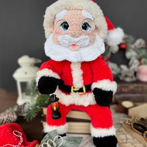 Pode incluir: Boneca de croch&ecirc; feita &agrave; m&atilde;o do Papai Noel, vestindo um terno vermelho com acabamento branco, luvas e botas pretas e um cinto preto. A boneca tem barba branca, &oacute;culos e um chap&eacute;u vermelho com acabamento de pele branca. Ela segura uma pequena lanterna.