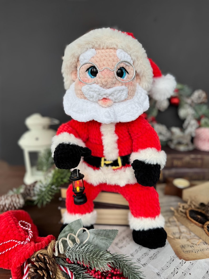Pode incluir: Boneca de croch&ecirc; feita &agrave; m&atilde;o do Papai Noel, vestindo um terno vermelho com acabamento branco, luvas e botas pretas e um cinto preto. A boneca tem barba branca, &oacute;culos e um chap&eacute;u vermelho com acabamento de pele branca. Ela segura uma pequena lanterna.