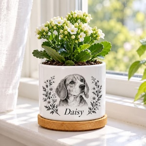 May include: A white ceramic cylindrical planter with a bamboo saucer base, featuring a black and white illustration of a beagle dog surrounded by floral sprigs and the name Daisy printed in black text, containing a white flowering plant.