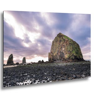 May include: A landscape photograph of a large rock formation on a beach under a cloudy sky. The rock is covered in green vegetation. The foreground is a dark, rocky beach. The sky is a mix of purple and gray.