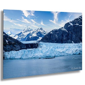 Può includere: Fotografia di un paesaggio di un ghiacciaio e montagne sotto un cielo blu con nuvole. Il ghiacciaio è di colore azzurro chiaro e le montagne sono di colore grigio scuro con cime innevate. L'acqua in primo piano è di colore blu scuro.