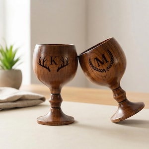 Peut inclure: Deux verres à vin en bois de couleur marron foncé. L'un porte la lettre "K" avec des bois de cerf, l'autre la lettre "M" dans une couronne de laurier. Les verres sont posés sur une surface claire.
