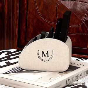 Könnte beinhalten: Beiger Fernbedienungshalter aus Stoff mit dem Buchstaben "M" in einem Lorbeerkranz. Der Halter enthält mehrere schwarze Fernbedienungen. Der Halter steht auf einem Stapel Bücher.