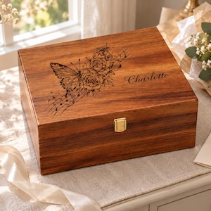 Könnte beinhalten: Eine rechteckige Erinnerungsbox aus Holz mit dunkelbraunem Finish und einem goldfarbenen Metallverschluss. Der Deckel zeigt eine Lasergravur eines Schmetterlings, Rosen und Noten sowie den in Schreibschrift eingravierten Namen Charlotte.