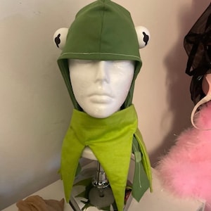 以下が含まれることがあります： 緑色のカエルをテーマにしたフードと襟のセット。フードには、黒い瞳の大きな白い目と尖った上部があります。襟は、尖った端を持つ明るい緑色です。コスチュームはマネキンの頭に表示されています。