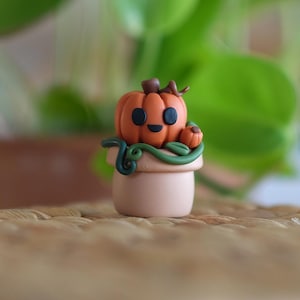 Puede incluir: Una figura en miniatura de calabaza naranja con una cara sonriente y un pequeño tallo, colocada sobre una pequeña maceta marrón claro. Enredaderas verdes envuelven la maceta. El fondo presenta follaje verde y una superficie tejida.