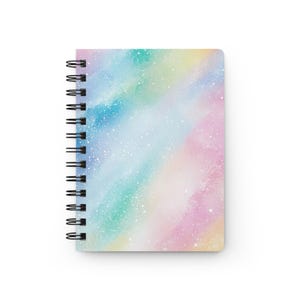 Op de afbeelding: Spiraalgebonden notitieboek met een kleurrijk, abstract ontwerp. De omslag heeft een aquarel-achtige mix van pastelkleuren, waaronder roze, blauw, groen en geel, met witte spikkels. Het notitieboek heeft een zwarte spiraalbinding.