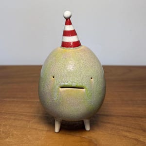 May include: A round, light green ceramic figurine with a speckled glaze, featuring two small eyes, a horizontal slit mouth, and two short white legs. A pointed party hat with red and white horizontal stripes sits on top of the figurine.