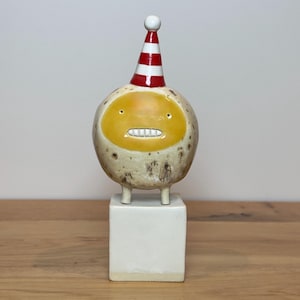May include: A ceramic figurine featuring a round, speckled beige body with a yellow face, small eyes, and a row of white teeth. The figure wears a red and white striped party hat with a white pom-pom on top and stands on a white square base.