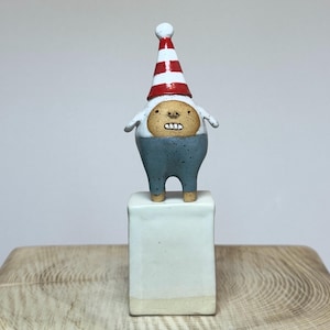 May include: A small ceramic figurine of a creature with a tan face, white arms, and blue legs, wearing a red and white striped pointed hat. The creature stands on a rectangular white ceramic block base with a smooth, glossy finish.