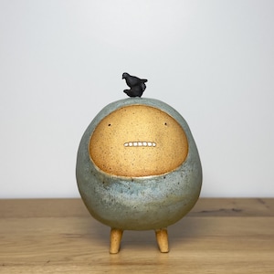 May include: A round, ceramic sculpture with a speckled, golden-tan face and a smooth, light blue-grey body. It features two small eyes, a horizontal line for a mouth with white teeth, and stands on two short legs with a small black bird on top.