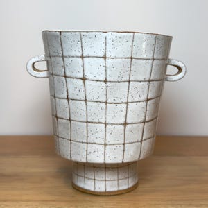 May include: A white ceramic vessel with a speckled finish and a brown grid pattern. The piece features a flared top, a narrow cylindrical base, and two small, rounded handles on opposite sides, showcasing a hand-crafted, textured appearance.