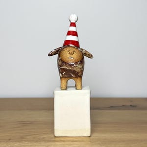 May include: A small ceramic figurine of a creature with a brown mottled body, two visible teeth, and a pointed party hat with red and white horizontal stripes, standing on top of a plain, rectangular, off-white ceramic block base.