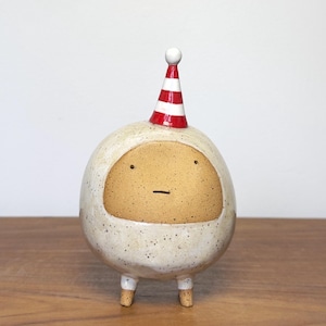 May include: A round ceramic figurine with a speckled off-white body and a smooth tan face. The figure features two small dark eyes, a simple horizontal line for a mouth, two short legs, and a pointed party hat with red and white horizontal stripes.