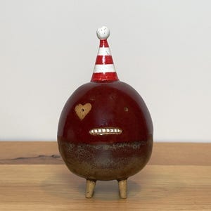 May include: A round ceramic figurine with a glossy dark red upper body and a textured brown base. It features two small legs, a heart-shaped eye, a simple dot eye, a row of white teeth, and a pointed party hat with red and white stripes.