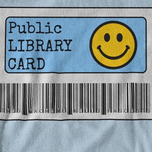 Peut inclure: T-shirt bleu clair avec un motif de carte de bibliothèque. Le motif de la carte comprend les mots "Public Library Card" en noir, un smiley jaune et un code-barres. Le texte "THIS MACHINE KILLS FASCISTS" est imprimé en dessous.