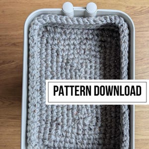 May include: A gray crocheted basket with a white rectangular label that reads "PATTERN DOWNLOAD". The basket has a sturdy, woven texture and a light gray rim. The basket is sitting on a light brown surface.