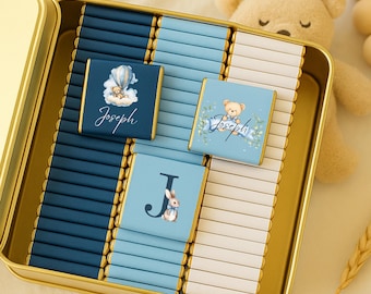 May include: A gold tin filled with chocolate bars, some wrapped in blue paper with illustrations of a hot air balloon, a teddy bear, and a bunny. The name "Joseph" is written on some of the wrappers. A stuffed teddy bear is in the background.
