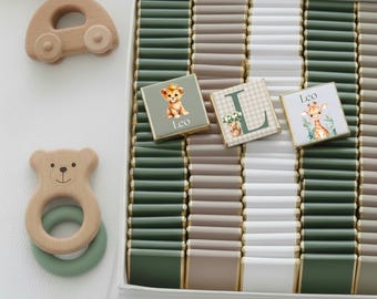 May include: A white box filled with rows of chocolates in green, white, and tan. Three decorated chocolates feature a lion, giraffe, and the letter "L". Wooden baby toys, including a car and bear-shaped teether, are on the left.