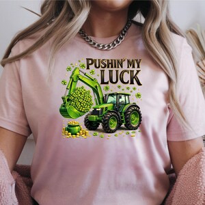 May include: A light pink t-shirt featuring a green tractor with a bucket full of green clovers and a pot of gold. The text "PUSHIN' MY LUCK" is above the tractor. The shirt is a crew neck style.