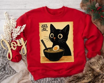 May include: Red sweatshirt featuring a black cat illustration with a bowl of ramen and the Japanese character for love. The cat has large yellow eyes and is set against a tan background. The sweatshirt is a crew neck.