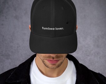May include: A black trucker hat featuring a mesh back and a solid front panel. The text "fumbwa lover." is embroidered in white on the center of the front panel. The hat has a structured crown and a flat brim, providing a classic silhouette.