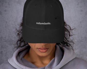 May include: A black trucker hat featuring a mesh back and a solid front panel. The word ndombolo. is embroidered in white lowercase letters across the center of the front panel. The hat has a curved brim and a structured crown design.
