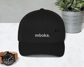 May include: A black baseball cap featuring the white embroidered text mboka. on the front panel. The cap has a structured crown, a curved brim, and a small black logo on the side. The design is simple, featuring a solid black fabric construction.