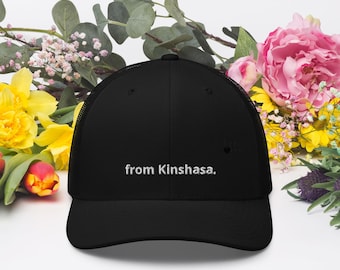 May include: A black trucker hat featuring a mesh back and a solid front panel. The text "from Kinshasa." is embroidered in white thread across the center of the front panel. The hat has a curved brim and a structured crown design.