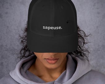 May include: A black trucker hat featuring a mesh back and a solid front panel. The word "sapeuse." is embroidered in white thread across the center of the front panel. The hat has a curved brim and a structured crown design with a button on top.