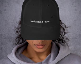 May include: A black trucker hat featuring a mesh back and a solid front panel. The text 'makemba lover.' is embroidered in white on the front center of the cap. The design includes a structured crown and a curved brim, providing a classic look.