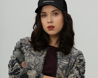 May include: A black baseball cap featuring the white text "mikate lover." printed on the front panel. The cap has a structured crown, a curved brim, and a solid black fabric construction. The design is centered on the front of the headwear piece.