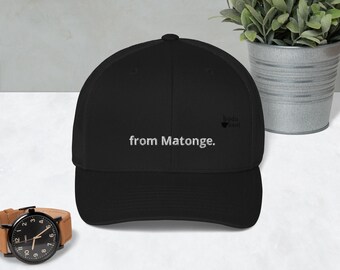 May include: A black baseball cap featuring the white embroidered text from Matonge. and a small black logo icon. The cap has a structured crown and a curved brim, with a classic six-panel design and a button on top, presenting a clean and simple look.