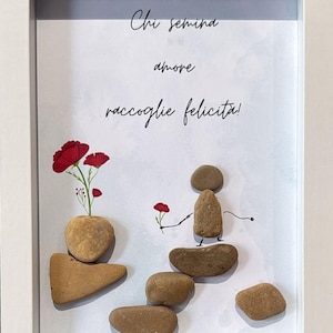 May include: Framed pebble art with a white border. The artwork shows a figure and flowers created from brown stones. Red flowers and the Italian text "Chi semina amore raccoglie felicità!" are also included in the design.