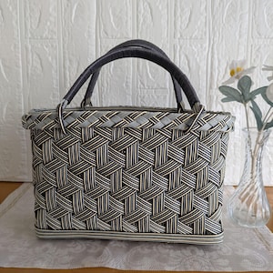May include: A rectangular woven basket with a dark gray handle and a complex geometric pattern in black, white, and light gray. The basket features a structured base and a reinforced rim, showcasing a tight, interlaced weave throughout the body.