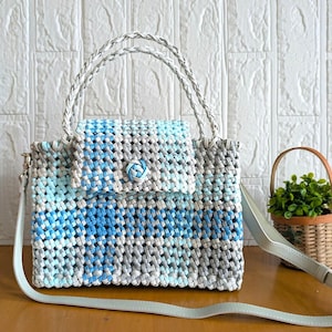 May include: A woven handbag featuring a blue, light blue, and grey checkered pattern. The bag has a braided handle, a flap closure secured with a central blue and white knot button, and a long, adjustable light blue shoulder strap.