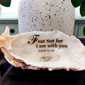 May include: A decorative seashell with a starfish accent. The shell is white with brown edges and features the engraved text "Fear Not for I am with you" from Isaiah 41:10. A light-colored starfish is attached to the shell.