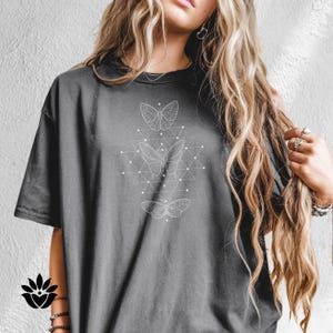 May include: Dark gray t-shirt featuring a white geometric butterfly design. The design includes three butterflies and a network of interconnected lines and dots. The shirt has a crew neck and short sleeves.