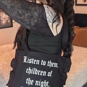 May include: A black tote bag featuring ruffled side edges and white text that reads: Listen to them, children of the night. What music they make. The bag has two shoulder straps and is constructed from a solid black fabric with a matte finish.