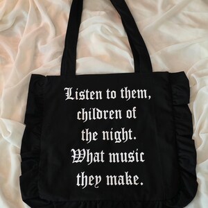 May include: A black tote bag featuring ruffled side and bottom edges. The front of the bag displays white text in a gothic font that reads: Listen to them, children of the night. What music they make. The bag has two long black shoulder straps.