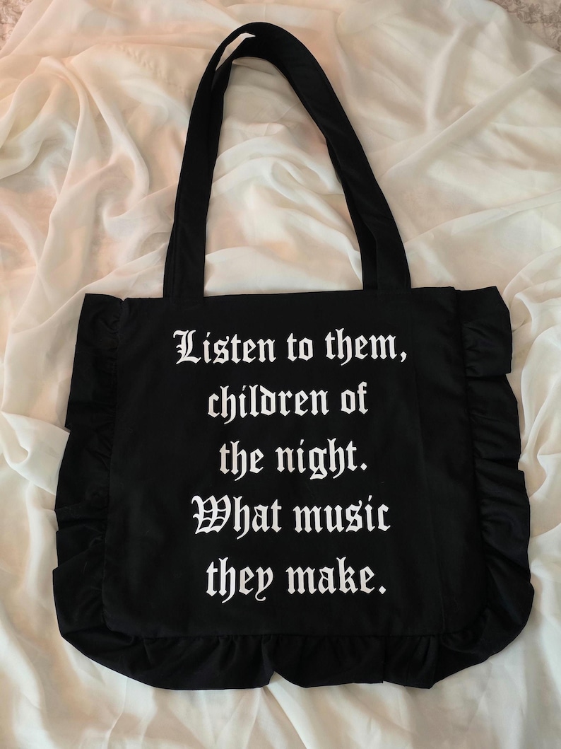 May include: A black tote bag featuring ruffled side and bottom edges. The front of the bag displays white text in a gothic font that reads: Listen to them, children of the night. What music they make. The bag has two long black shoulder straps.