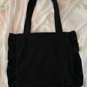 May include: A solid black tote bag featuring two long shoulder straps and a decorative ruffled trim along the left, right, and bottom edges. The bag has a square-shaped main compartment constructed from a smooth, dark fabric material.
