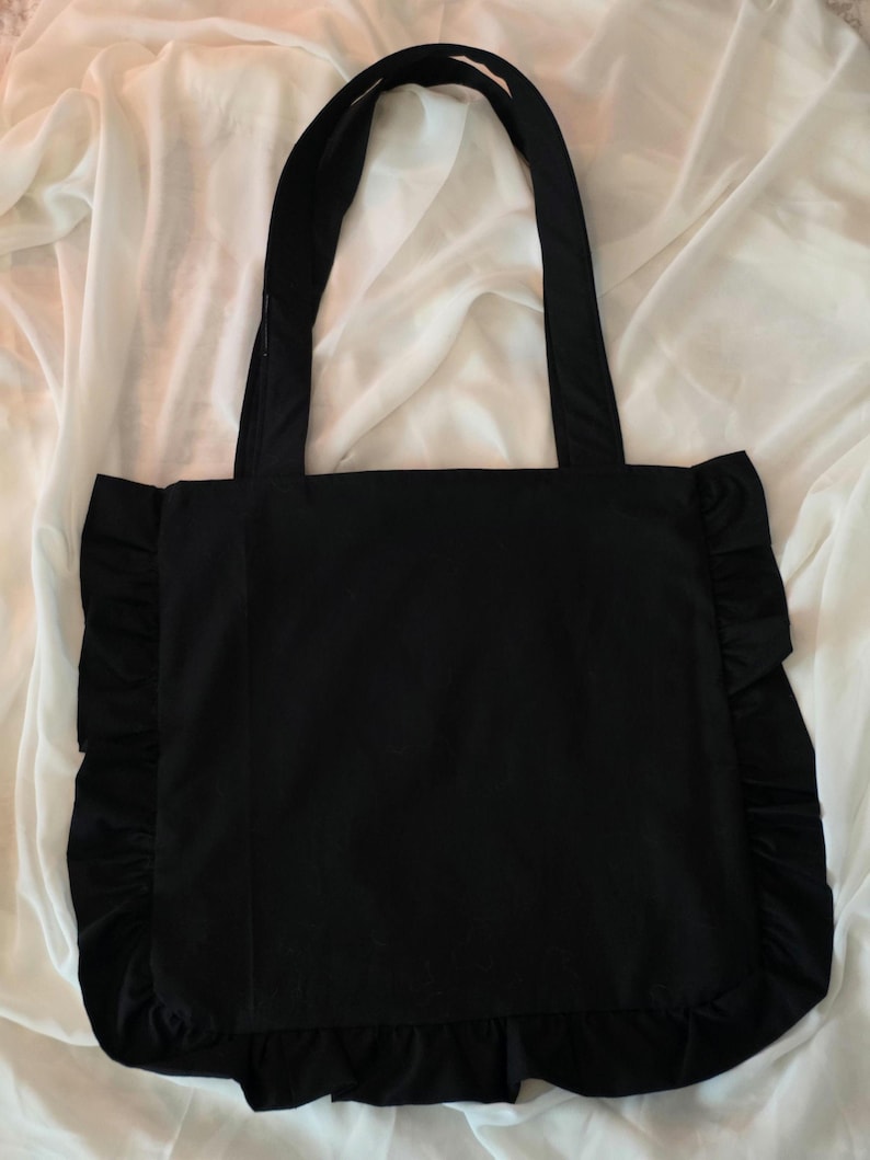 May include: A solid black tote bag featuring two long shoulder straps and a decorative ruffled trim along the left, right, and bottom edges. The bag has a square-shaped main compartment constructed from a smooth, dark fabric material.