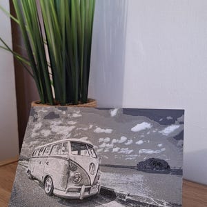 May include: Black and white image of a vintage Volkswagen bus parked on a road next to the ocean. The artwork is displayed on a wooden surface, with a green plant in the background.