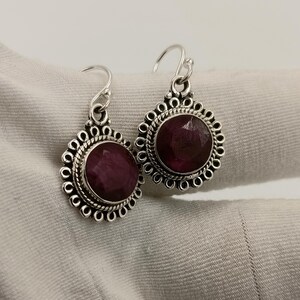 May include: A pair of silver earrings featuring large, round, deep red gemstones. Each earring has a decorative silver border with a scalloped design. The earrings have a hook closure. Jewellery.