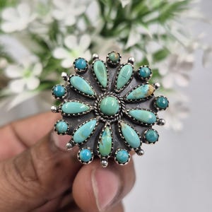 Könnte beinhalten: Ein silberner Ring mit einem türkisfarbenen Blumenmuster. Der Ring hat einen zentralen Türkisstein, umgeben von tropfenförmigen Türkissteinen und kleineren Türkisperlen. Der Ring ist vor einem verschwommenen weißen floralen Hintergrund.
