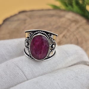 Peut inclure: Une bague en argent avec une grosse pierre précieuse ovale rouge foncé. La bague présente un design orné avec une bande texturée et des éléments décoratifs autour de la pierre précieuse. La bague est posée sur une surface blanche.